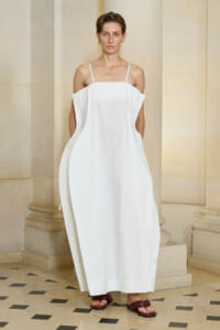 Model wearing a loose, white off-shoulder dress with thin straps, standing against a stone columned backdrop in a classical interior.