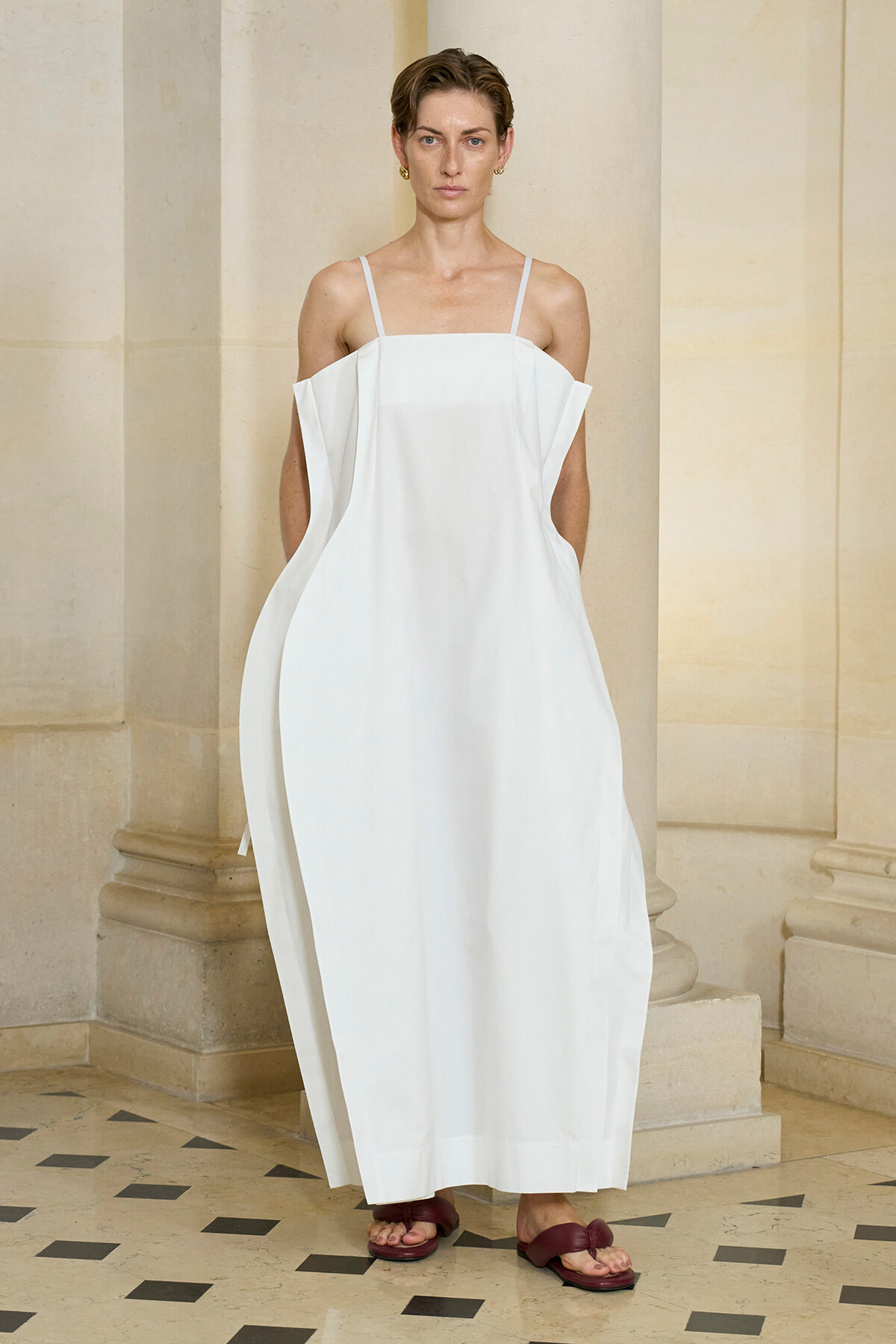 Model wearing a loose, white off-shoulder dress with thin straps, standing against a stone columned backdrop in a classical interior.