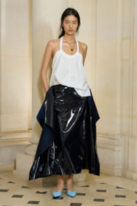 Model wearing a white halter top with a shiny black vinyl skirt and navy draped fabric, standing beside stone columns, wearing light blue flats and a gold pendant.