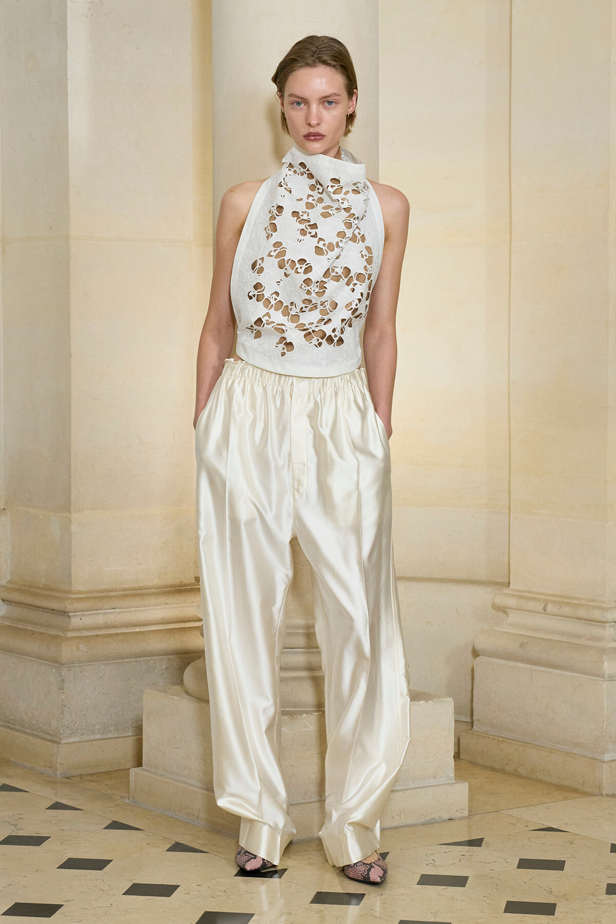 Model in a white lace high-collar halter top and ivory satin wide-leg trousers, standing against beige stone columns in a fashion shoot; pink snakeskin heels.