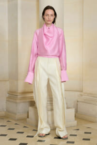 Model wearing a pink satin draped blouse with a scarf-like collar and cream wide-leg trousers, standing in a stone hallway.