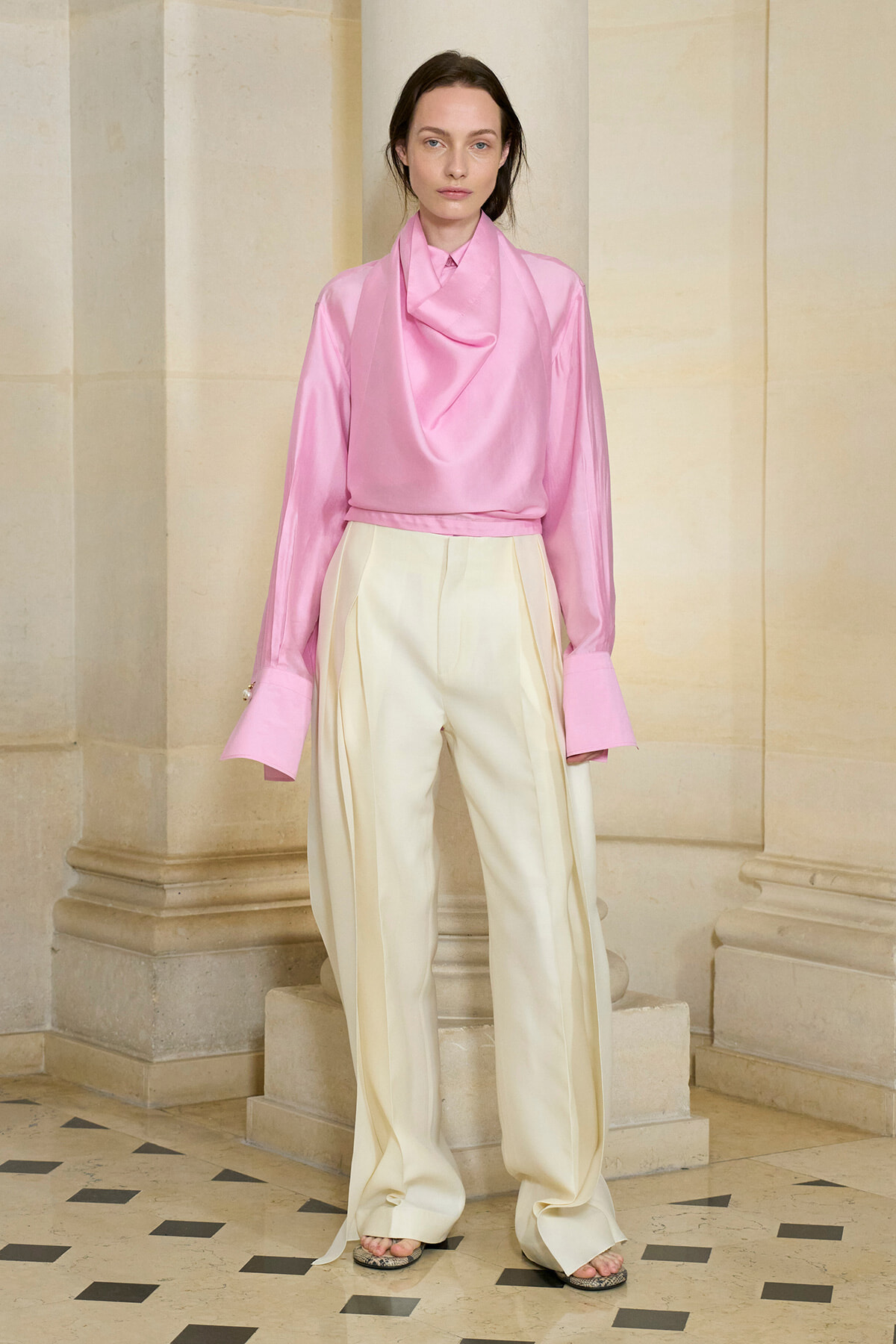 Model wearing a pink satin draped blouse with a scarf-like collar and cream wide-leg trousers, standing in a stone hallway.