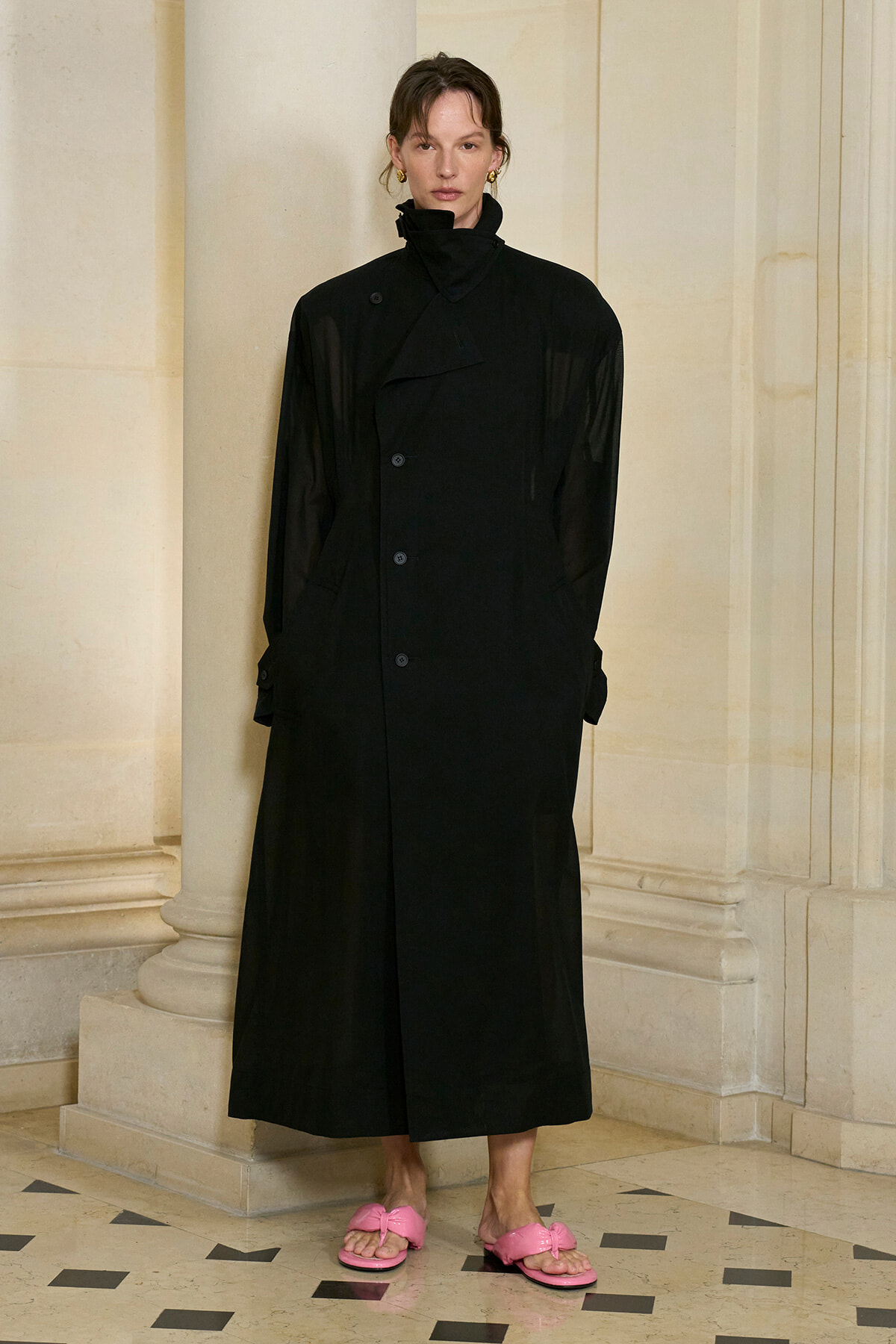Person wearing a long black coat with a high collar, standing against pale stone columns; pink slide sandals visible.