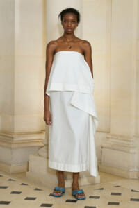 Model wearing a strapless white midi dress with a layered draped bodice, standing in a stone-colonnade setting with gold jewelry and blue flat sandals.