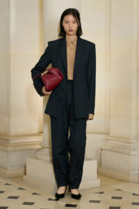 Model wearing an oversized black suit with a tan button-down shirt, holding a red handbag, standing by stone columns in a hallway