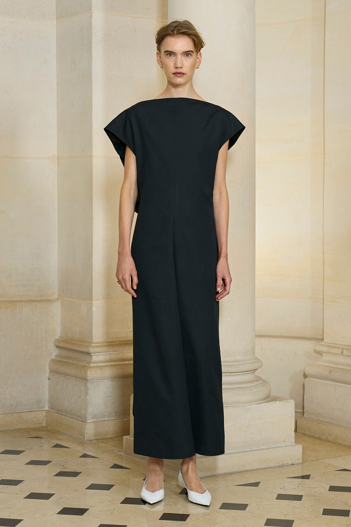 Woman in a minimalist black wide-leg jumpsuit with short, angular sleeves, standing in a stone colonnade.