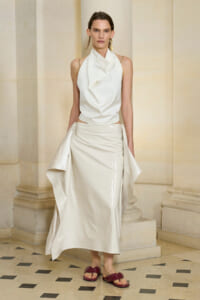 Model wearing an ivory draped sleeveless top with a glossy midi skirt and burgundy sandals, posed in a stone foyer setting.