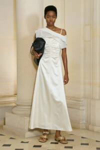 Model wearing a white off-shoulder satin gown with a ruched bodice, standing beside columns and carrying a black handbag.
