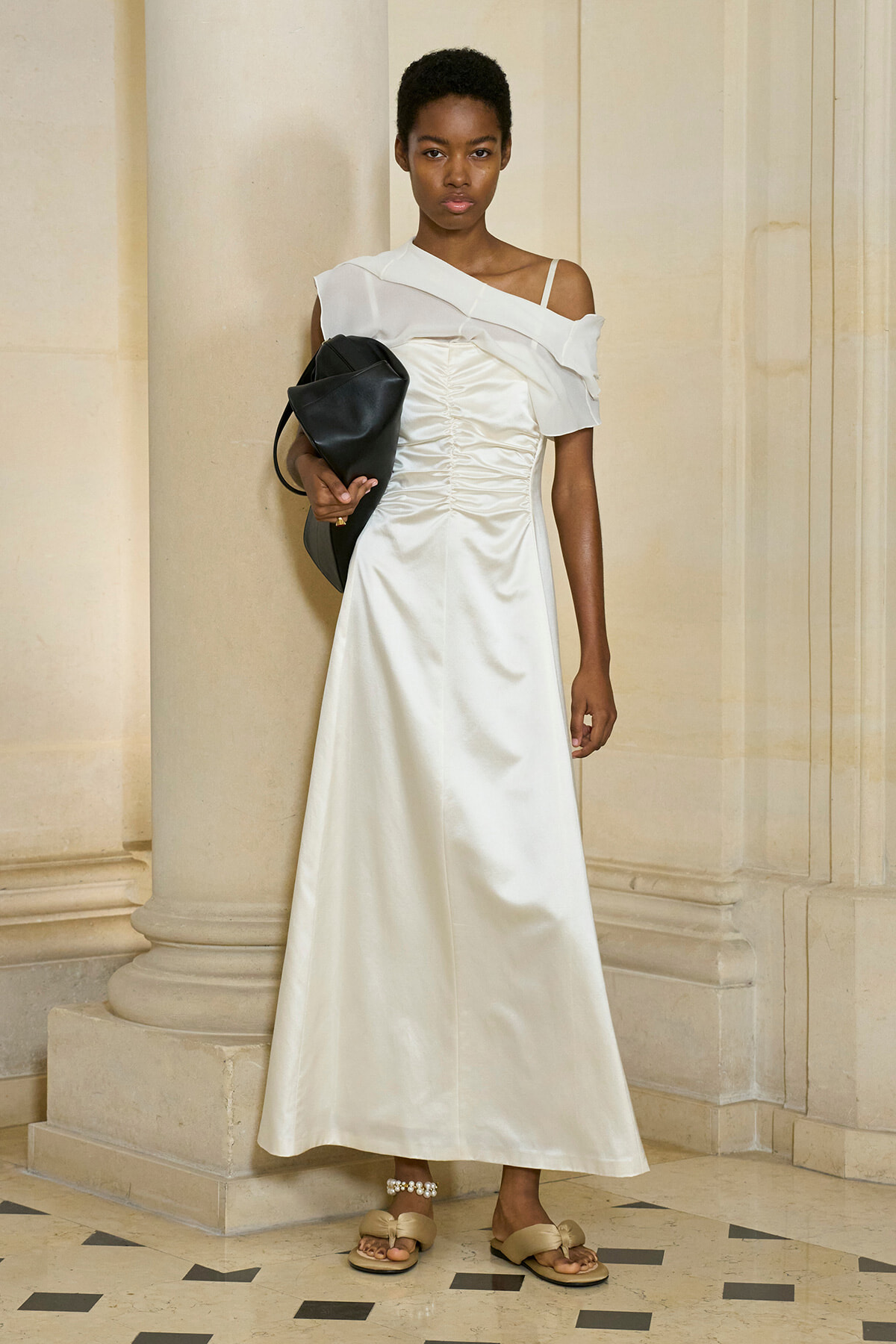 Model wearing a white off-shoulder satin gown with a ruched bodice, standing beside columns and carrying a black handbag.