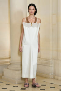 Model standing in a stone hallway, wearing a white lace-trim slip dress with thin straps and a brown underlayer, accessorized with a gold pendant necklace and snakeskin sandals.