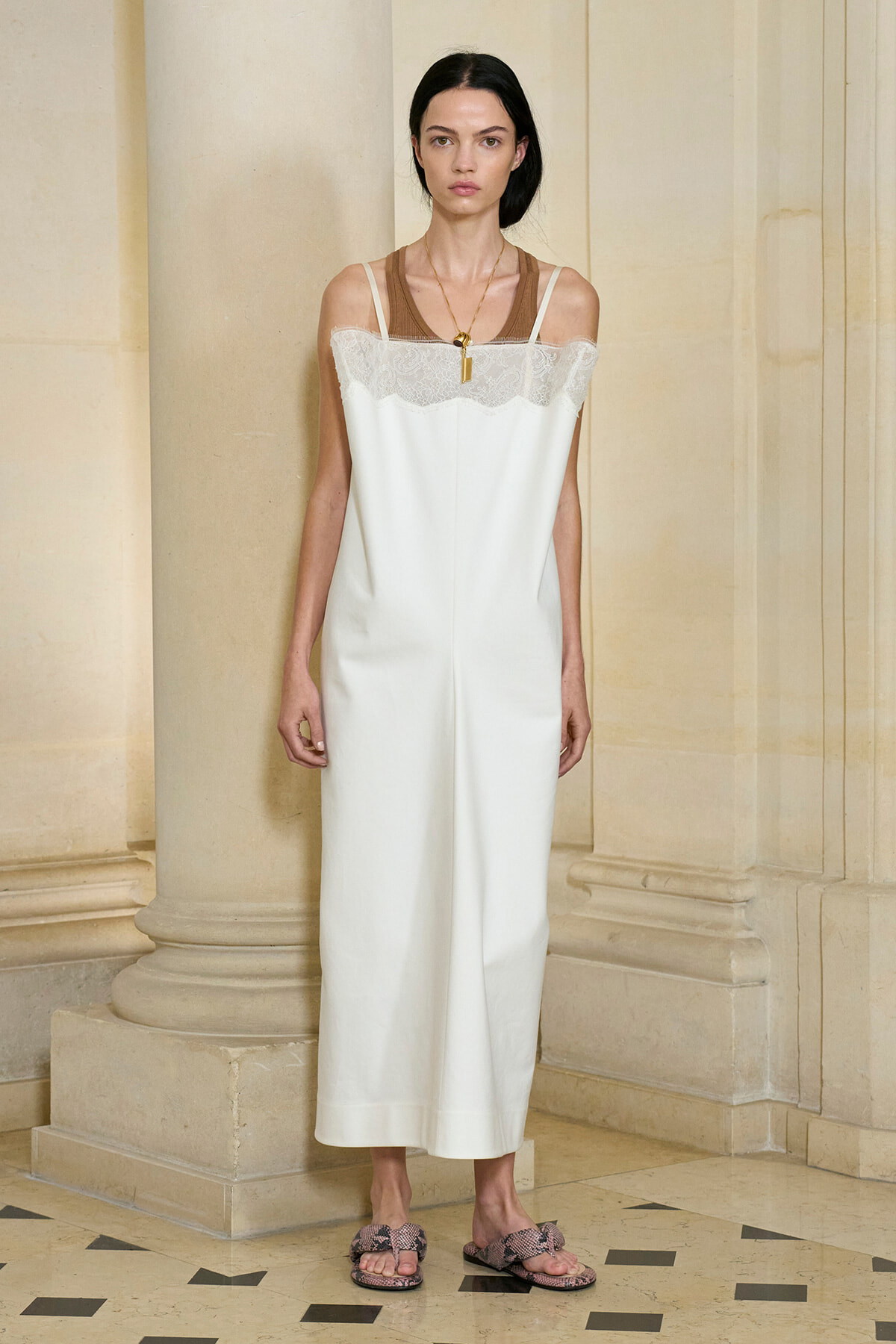 Model standing in a stone hallway, wearing a white lace-trim slip dress with thin straps and a brown underlayer, accessorized with a gold pendant necklace and snakeskin sandals.