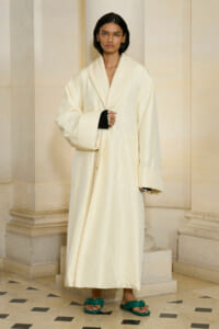 Woman in a long, oversized cream coat posed against stone columns, wearing green sandals.