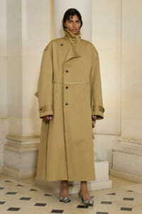 Woman wearing a tan oversized trench coat with a high collar, standing in a light stone hallway with columns.