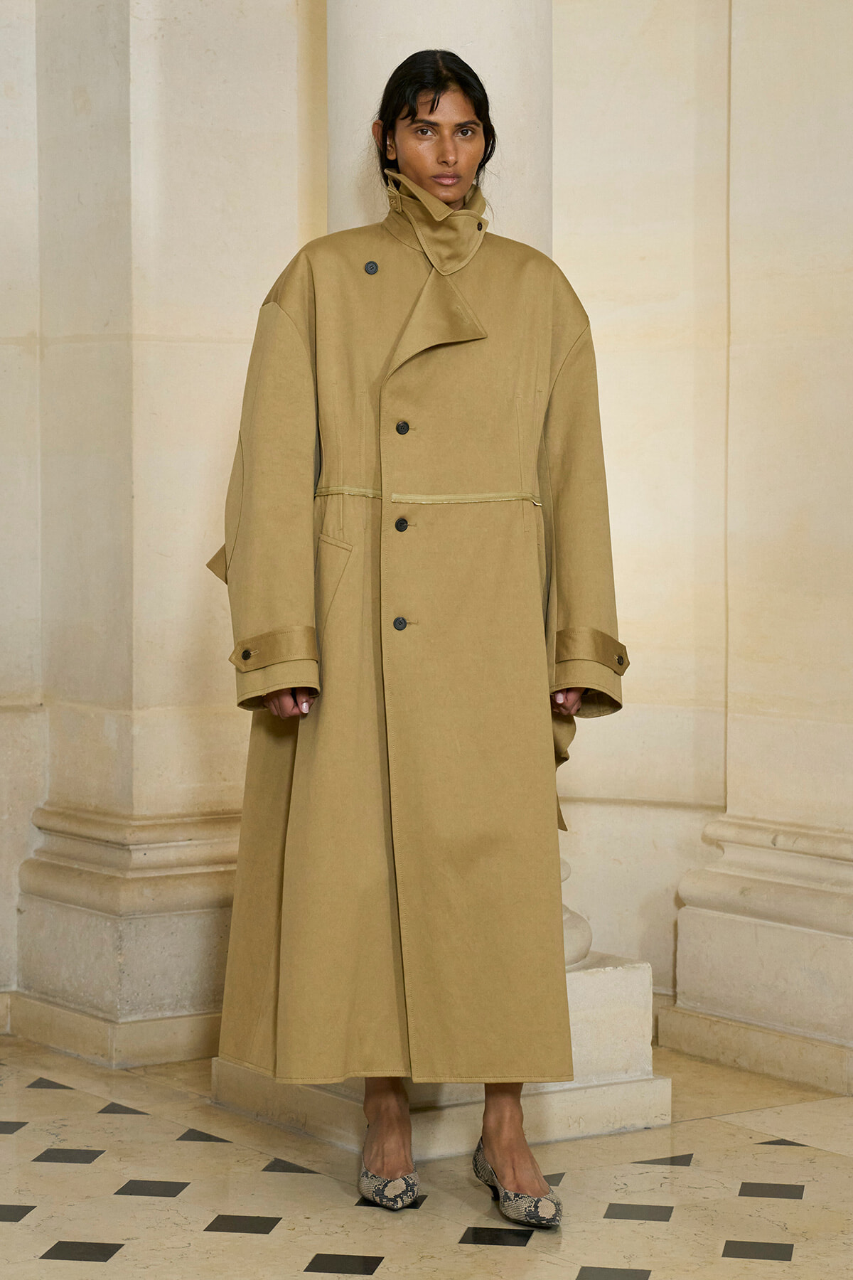 Woman wearing a tan oversized trench coat with a high collar, standing in a light stone hallway with columns.