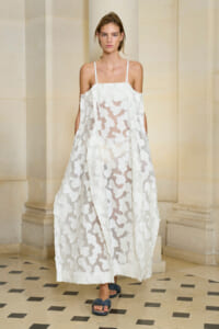 Model wearing a white sheer gown with large floral appliqués and thin shoulder straps, standing in a light stone corridor.