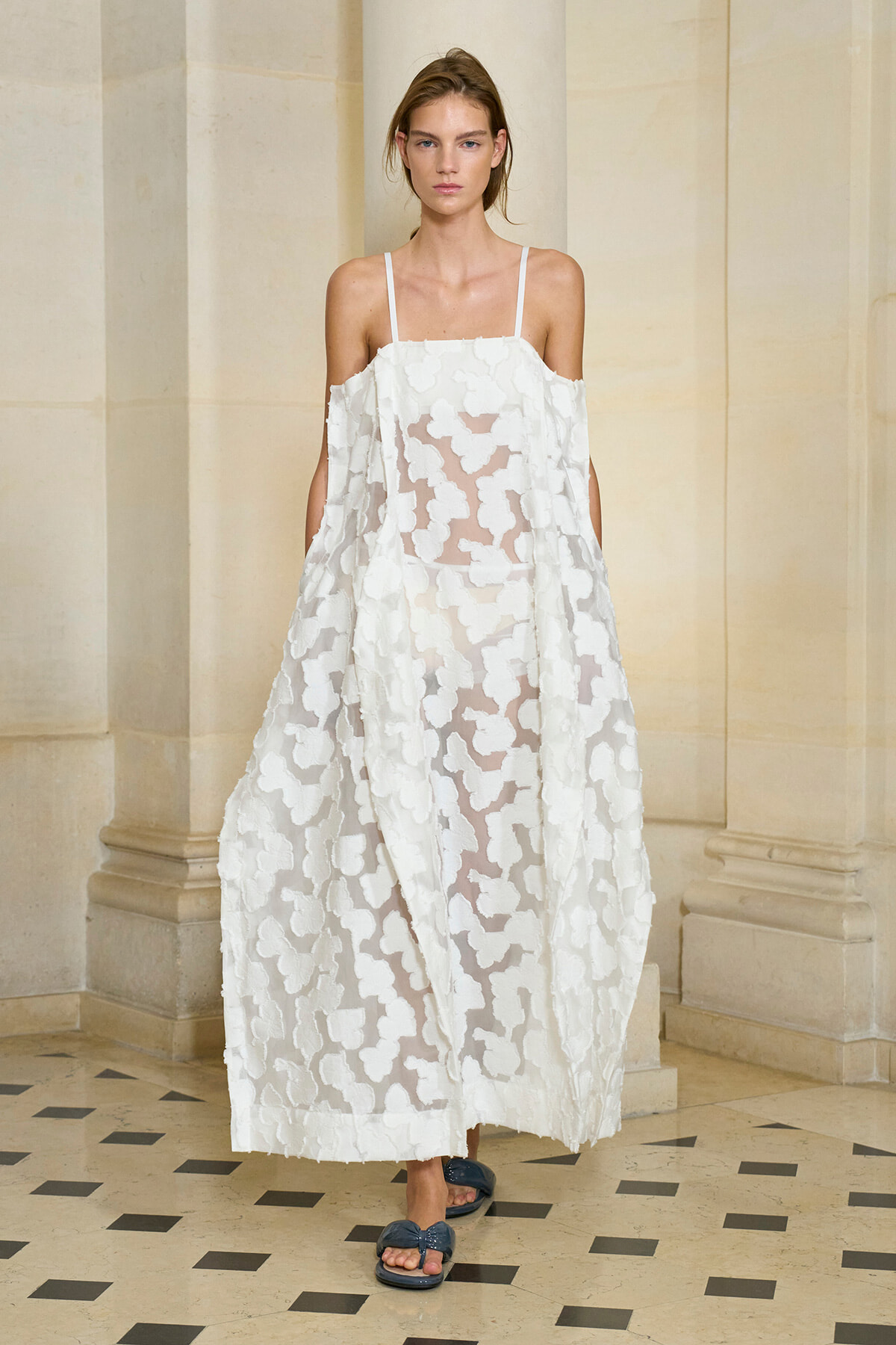 Model wearing a white sheer gown with large floral appliqués and thin shoulder straps, standing in a light stone corridor.