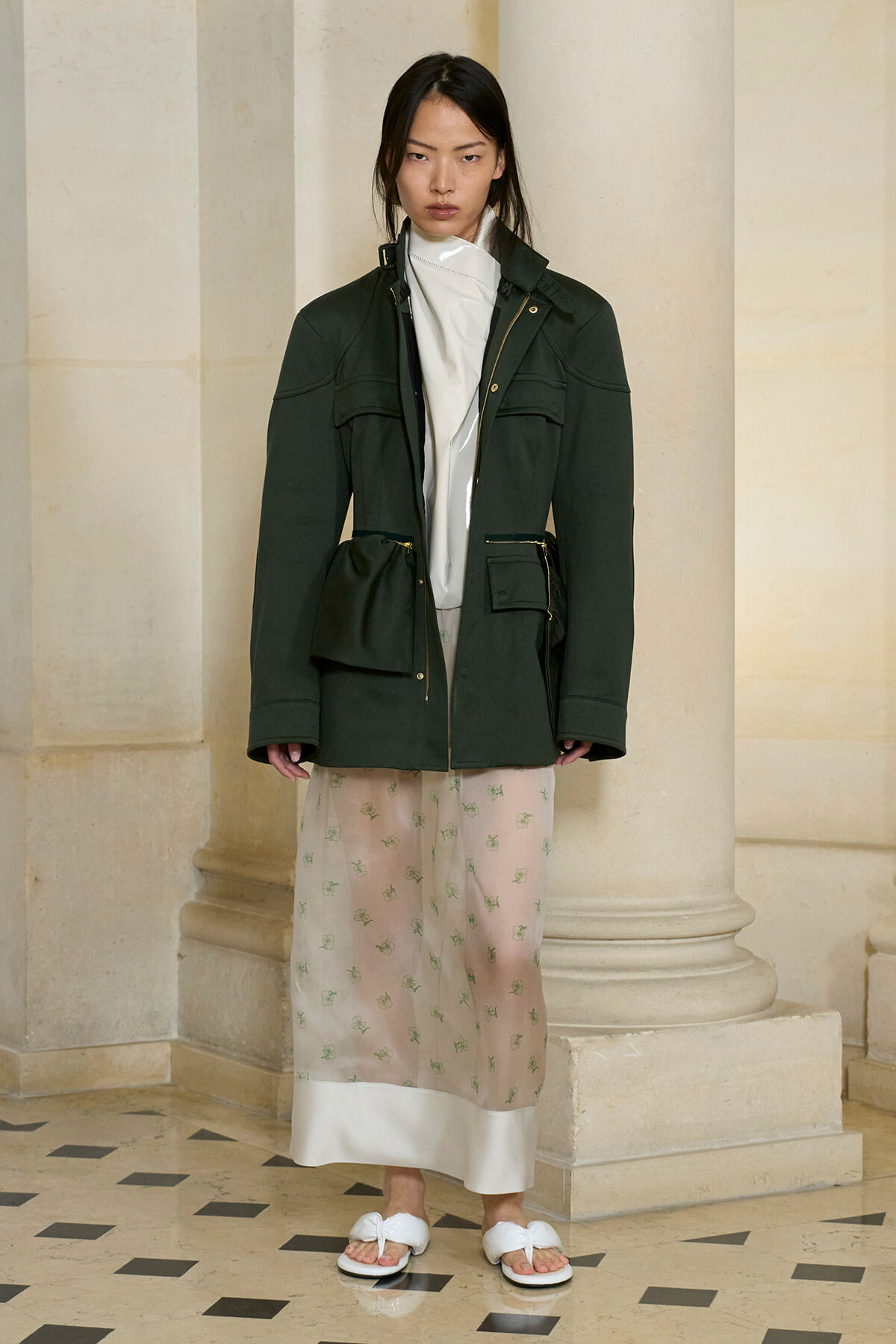Model standing beside stone columns in a dark green parka with gold zippers, white top, pale pink floral midi skirt, and white sandals.