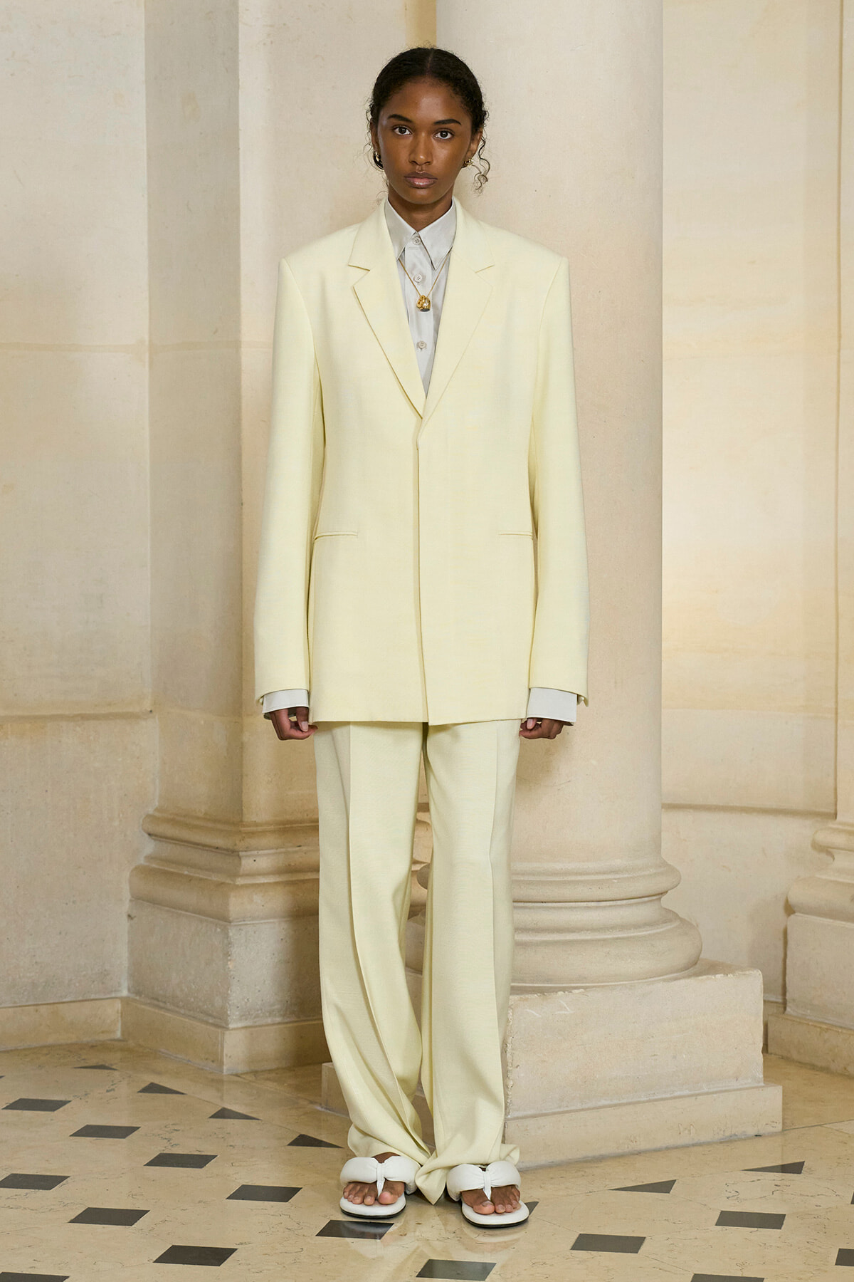 Woman in a pale yellow oversized suit (blazer and wide-leg pants) with a white shirt, standing in a marble-columned interior.