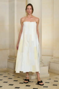 Model wearing a strapless cream patchwork midi dress, standing in a stone-columned interior.