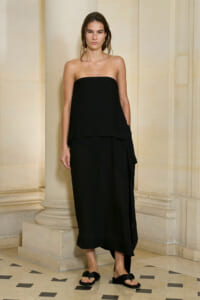 Woman wearing a strapless black midi dress and black slide sandals stands against a columned stone wall.