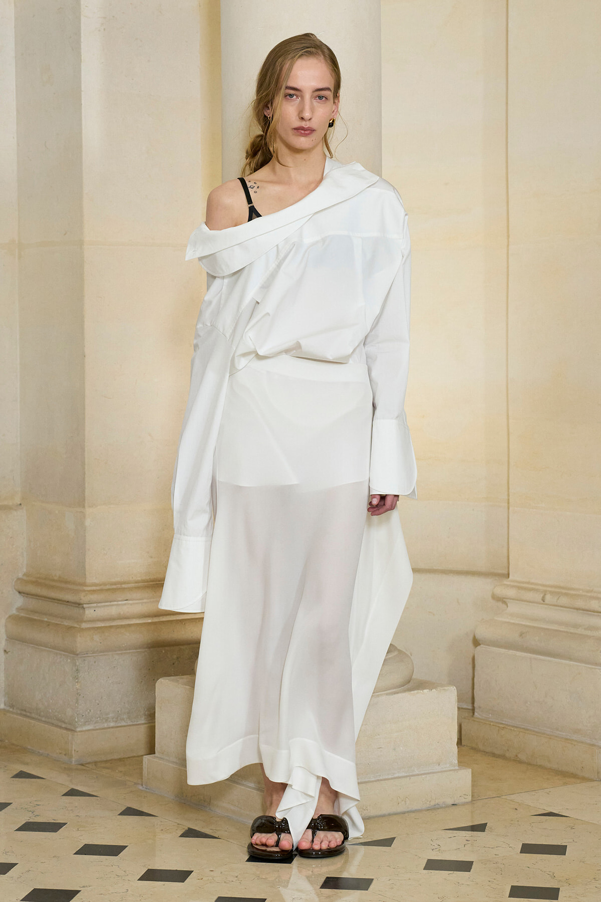 Model wearing an off-shoulder oversized white dress with draped layers, standing beside limestone columns in a stone hallway, wearing black sandals.