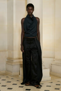Fashion model in an all-black outfit with a draped sleeveless top and wide-leg trousers, posing against light stone columns.
