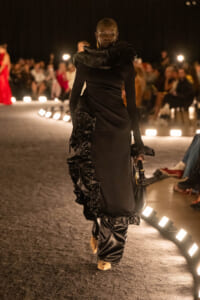 Runway model in a sleek black gown with dramatic ruched ruffles, walking a lit catwalk as an attentive audience watches.
