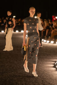 Model walks a nighttime fashion runway in a gray knit dress with circular cutouts, carrying a black handbag and gold accents.