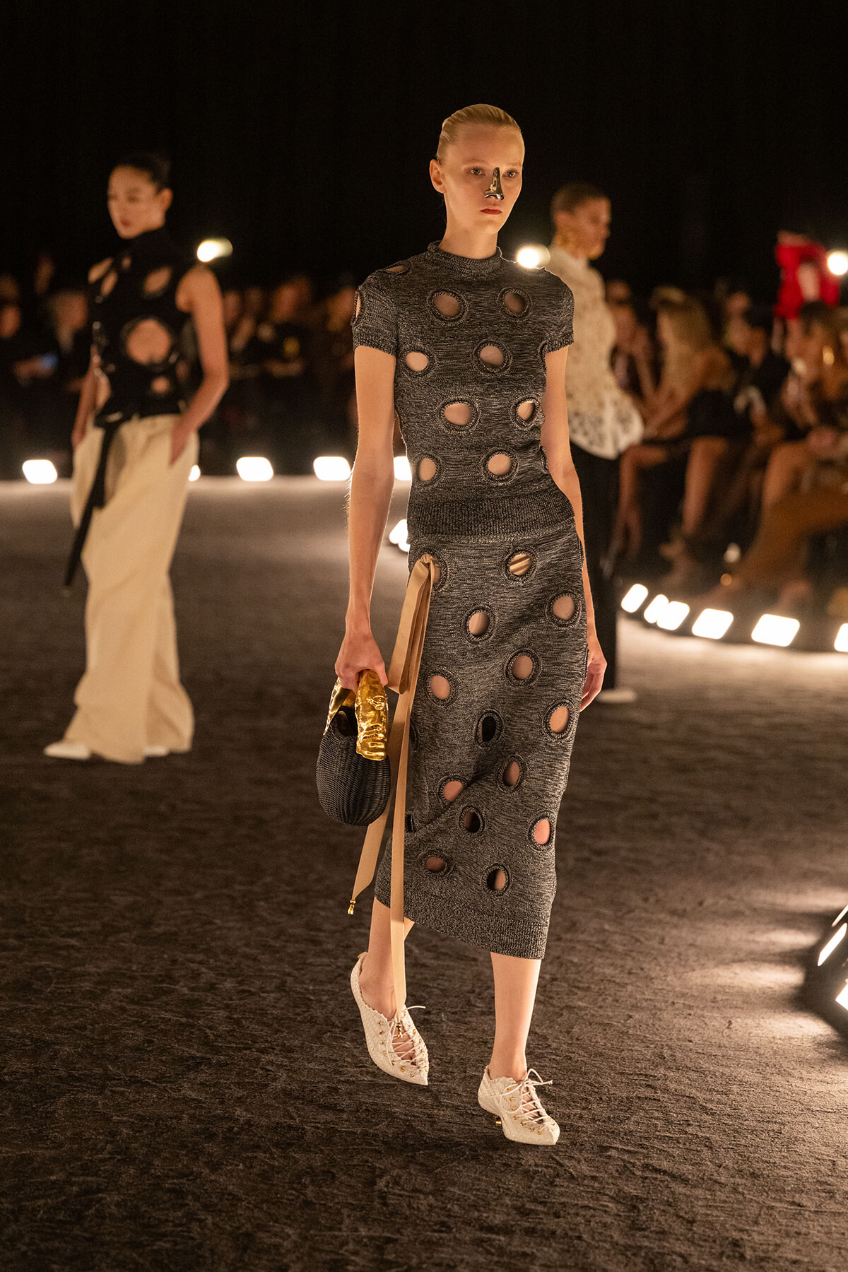 Model walks a nighttime fashion runway in a gray knit dress with circular cutouts, carrying a black handbag and gold accents.