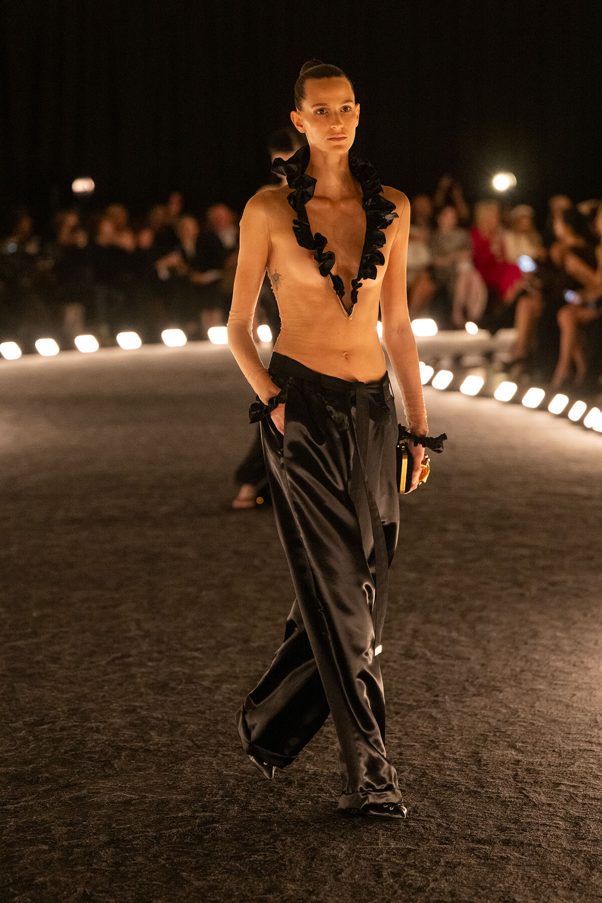 Female model walks a fashion runway at night wearing black satin pants and a dramatic ruffled necklace.