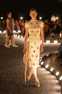 Model on a runway wearing a cream knit dress with large circular cutouts, carrying a beige bag, at a nighttime fashion show.