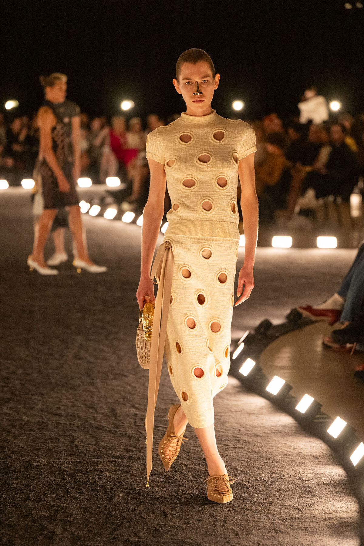 Model on a runway wearing a cream knit dress with large circular cutouts, carrying a beige bag, at a nighttime fashion show.