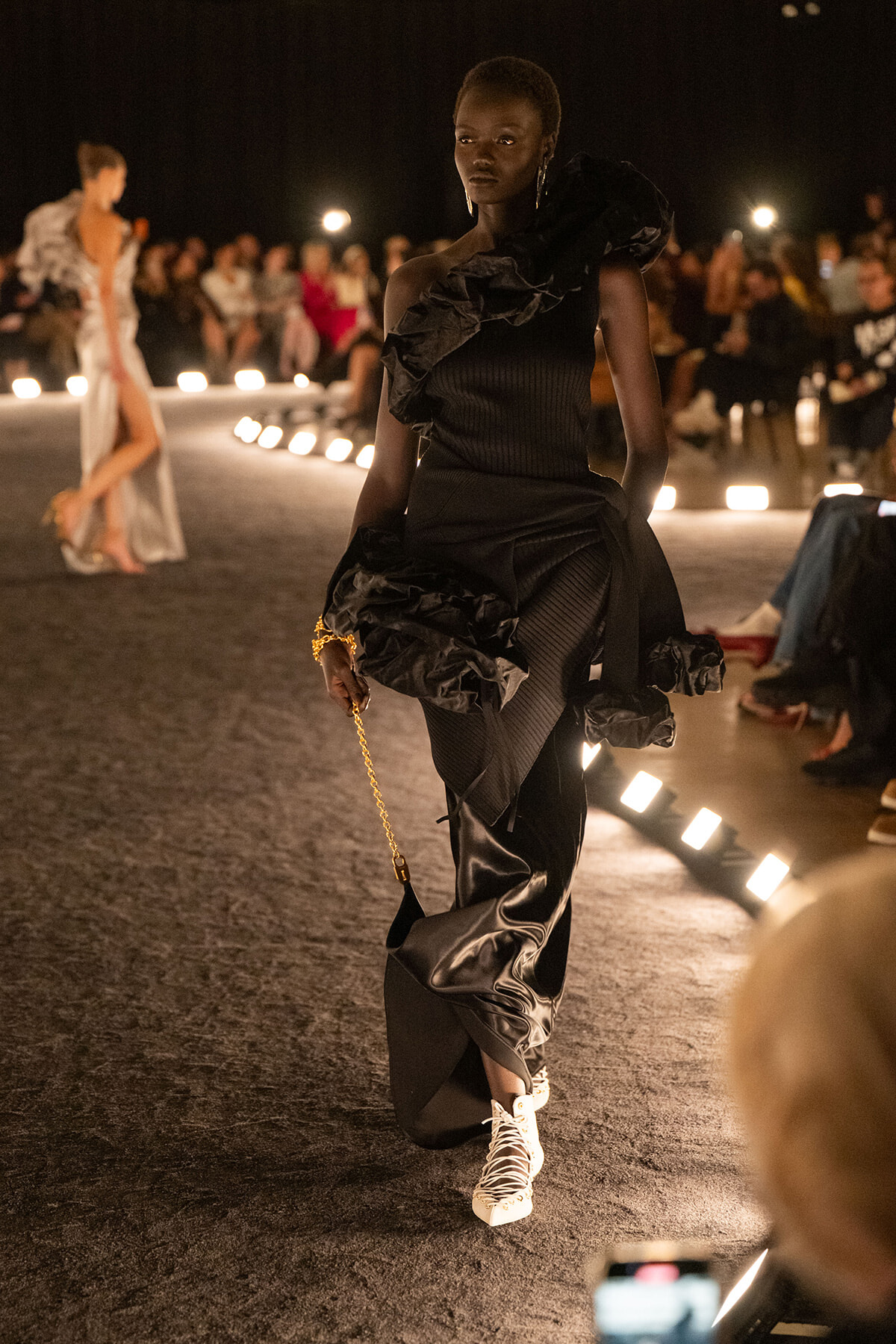 Model on a dimly lit runway in a black ruffle-shoulder dress, carrying a gold chain purse and wearing white sneakers.