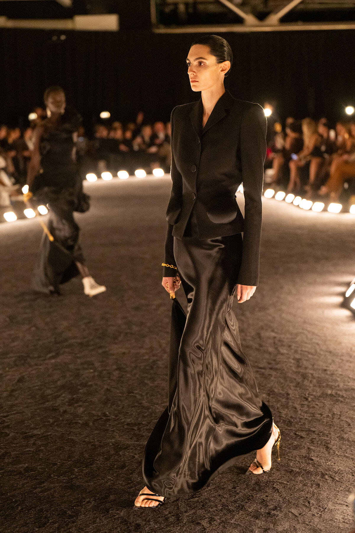 Model walking a fashion runway in a black tailored blazer and long satin skirt, with an audience in the background on dimly lit set.