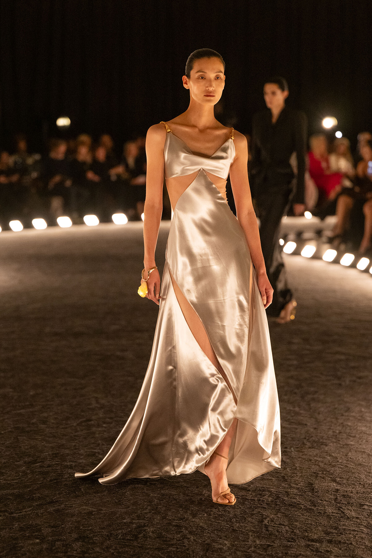 Model walks a silver satin evening gown with cutouts and thigh-high slit on a runway at night, audience in background.