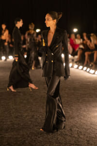Model walks the runway wearing a black double-breasted suit with gold buttons, slicked hair in a bun, and gloved hand accessories.