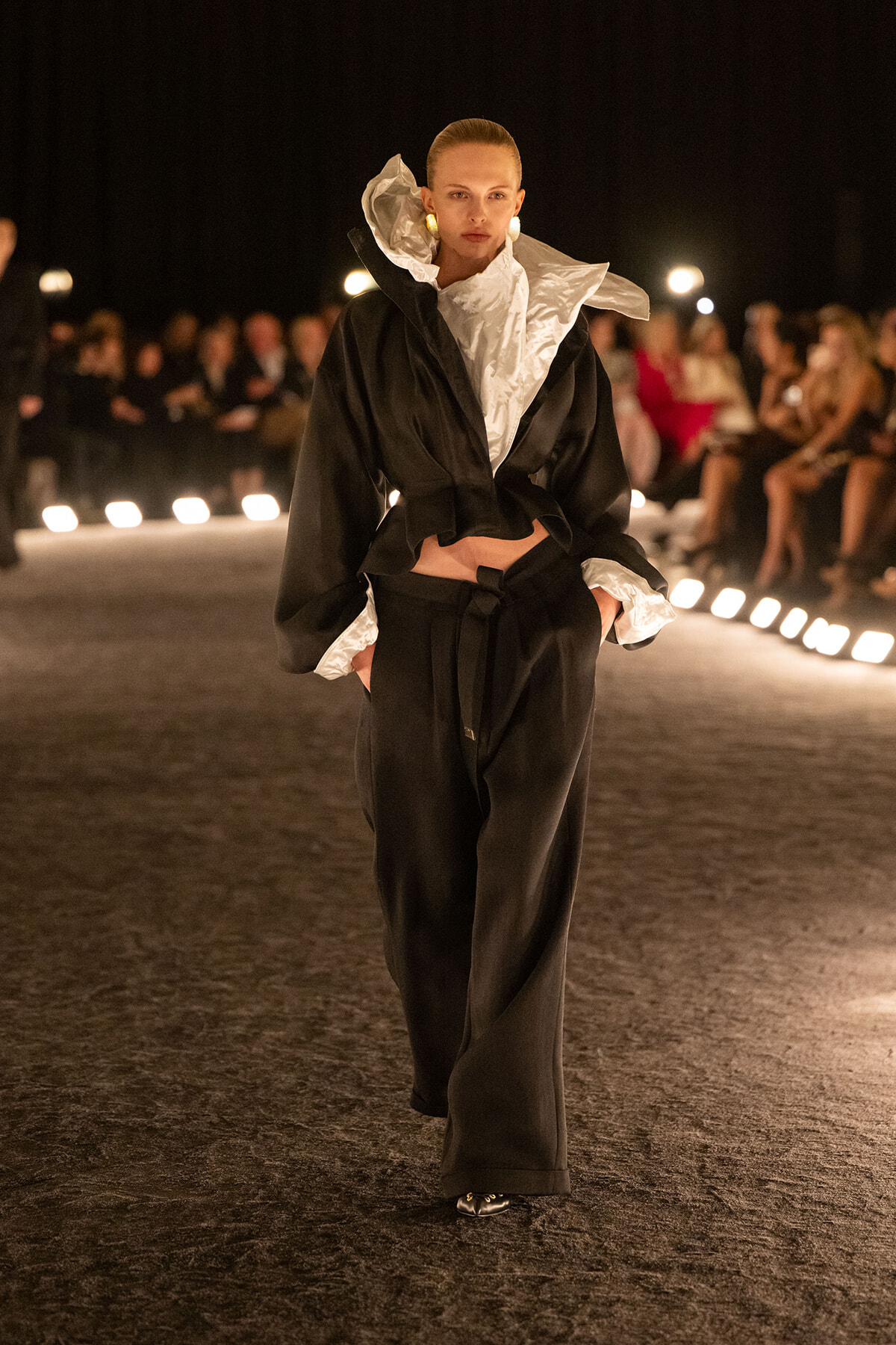 Model walks a night runway in a black ensemble with white ruffled collar and cuffs, hands in pockets, audience in background