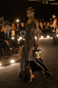 Model walking a runway in a black satin gown with sheer panels, gold headpiece and jewelry, carrying a black handbag with gold charms.