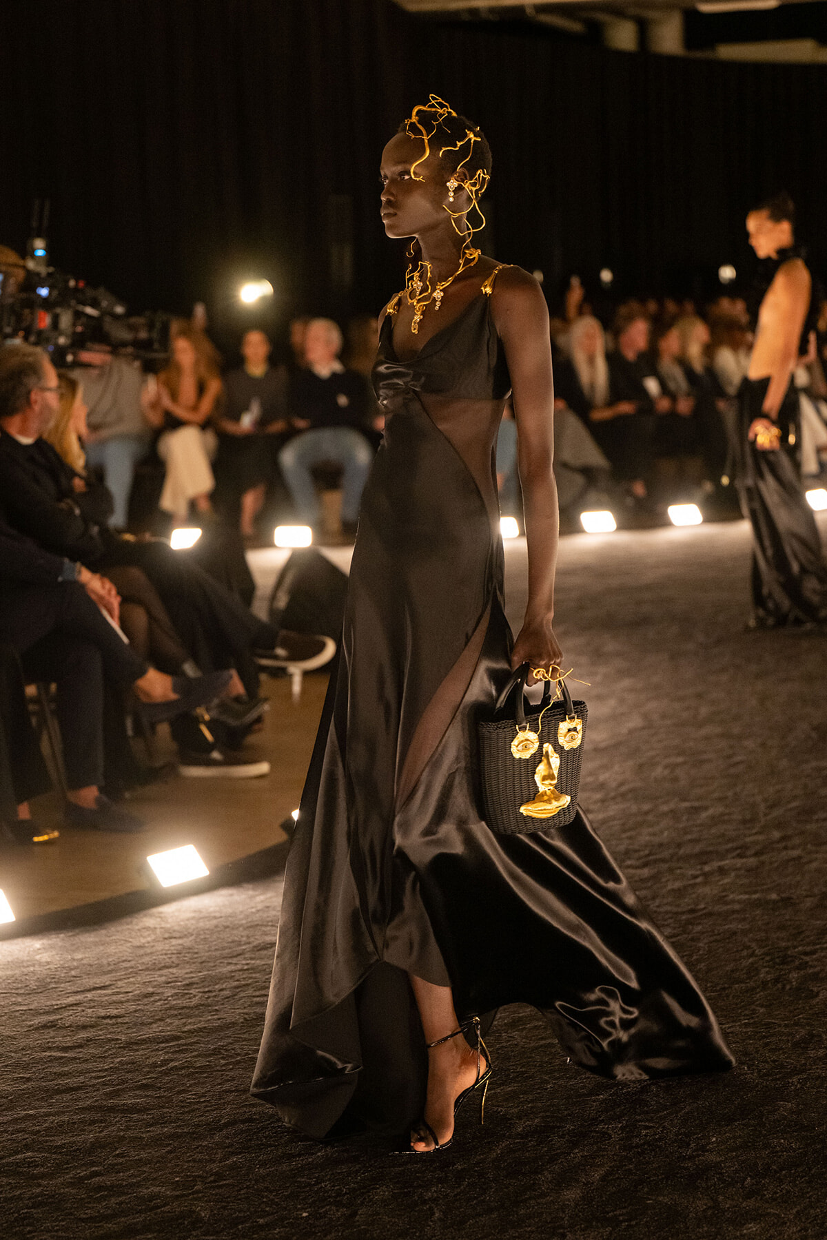 Model walking a runway in a black satin gown with sheer panels, gold headpiece and jewelry, carrying a black handbag with gold charms.