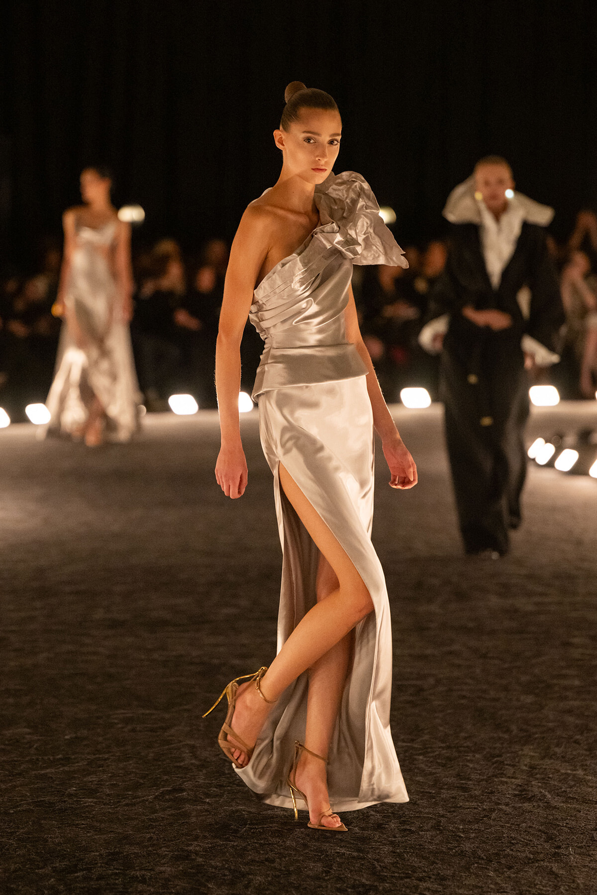 Model walks a silver one-shoulder gown with a thigh-high slit on a runway.
