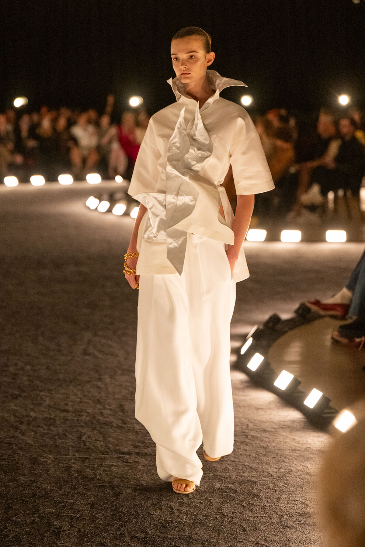 Model in an all-white avant-garde outfit with an oversized collar and sculptural scarf walking a runway, audience blurred in the background.