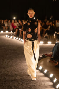 Model walks a dimly lit runway in a black circular-cutout top and cream wide-leg pants with a black belt, audience in background.