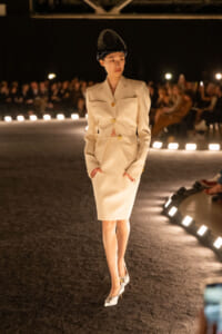 Female model walks a runway wearing a cream tailored blazer and skirt with gold buttons and a black decorative hat on her head.