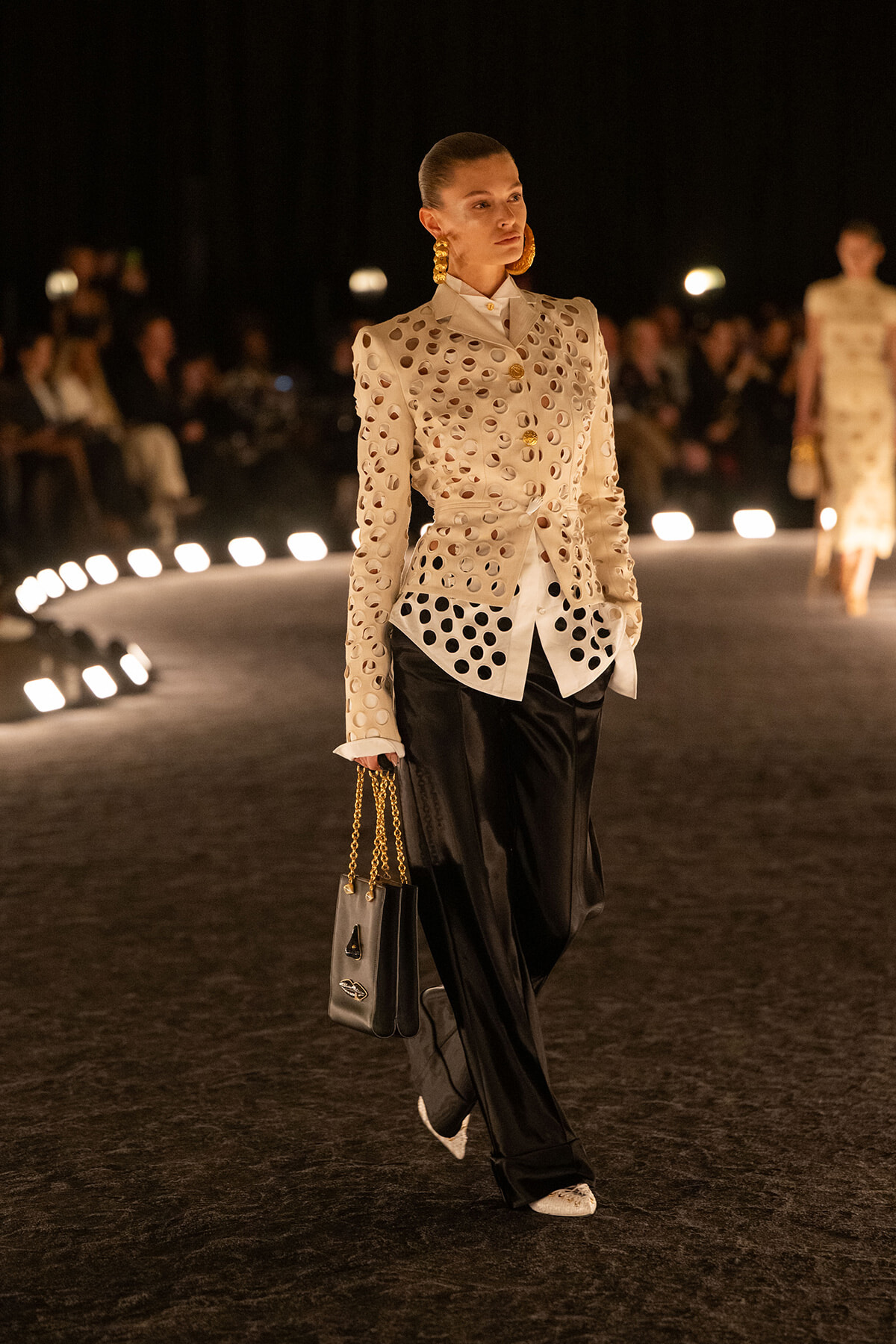 Model walking a runway at night in a perforated beige blazer with circular cutouts, black wide-leg trousers, white shoes, gold hoops, and a small black handbag with a gold chain.
