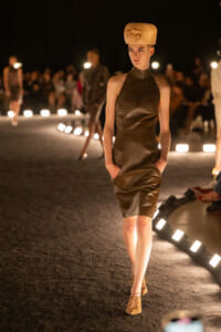 Model walks a fashion runway wearing a brown leather sheath dress with studded cups and tan lace-up shoes, against warm lighting.