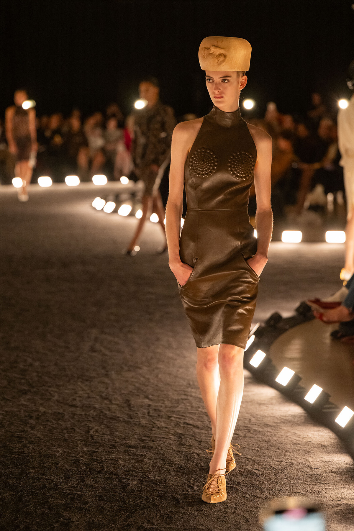 Model walks a fashion runway wearing a brown leather sheath dress with studded cups and tan lace-up shoes, against warm lighting.