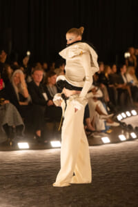 Model on a runway in an avant-garde white ensemble with a large sculptural collar and wide-legged pants; audience in the background.