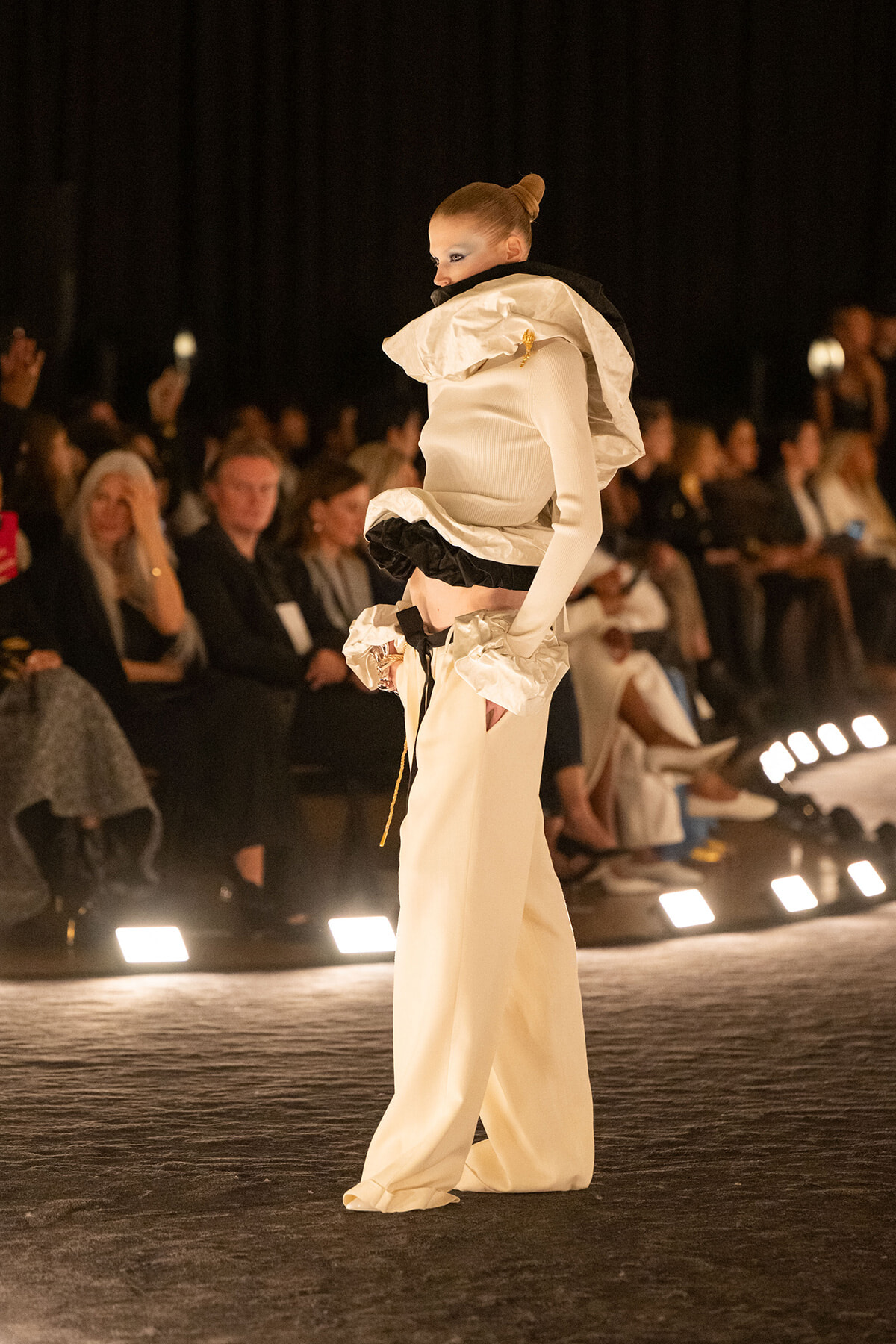 Model on a runway in an avant-garde white ensemble with a large sculptural collar and wide-legged pants; audience in the background.