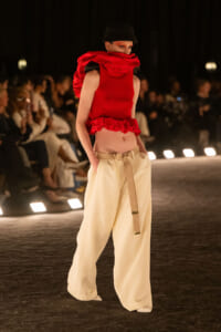 Runway model in a red sleeveless top with ruffled scarf and beige wide-leg pants, wearing a black beanie; audience blurred in the background.
