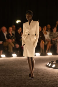 Model walks down a dimly lit runway in a cream structured suit, hands in pockets, audience blurred in the background.